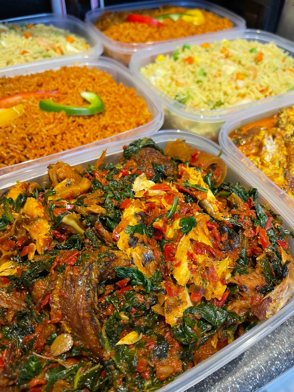 Tray of efo riro spinach stew with fish and meat beside trays of jollof and fried rice